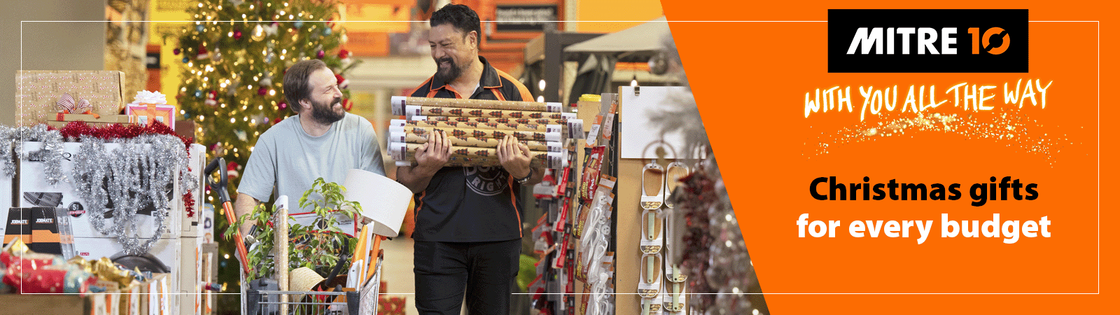 A customer and Mitre 10 staff member walk through a Mitre 10 store carrying wrapping paper and a trolley full of product. Accompanied by a heading "Loads of Christmas gift ideas to suit your budget"